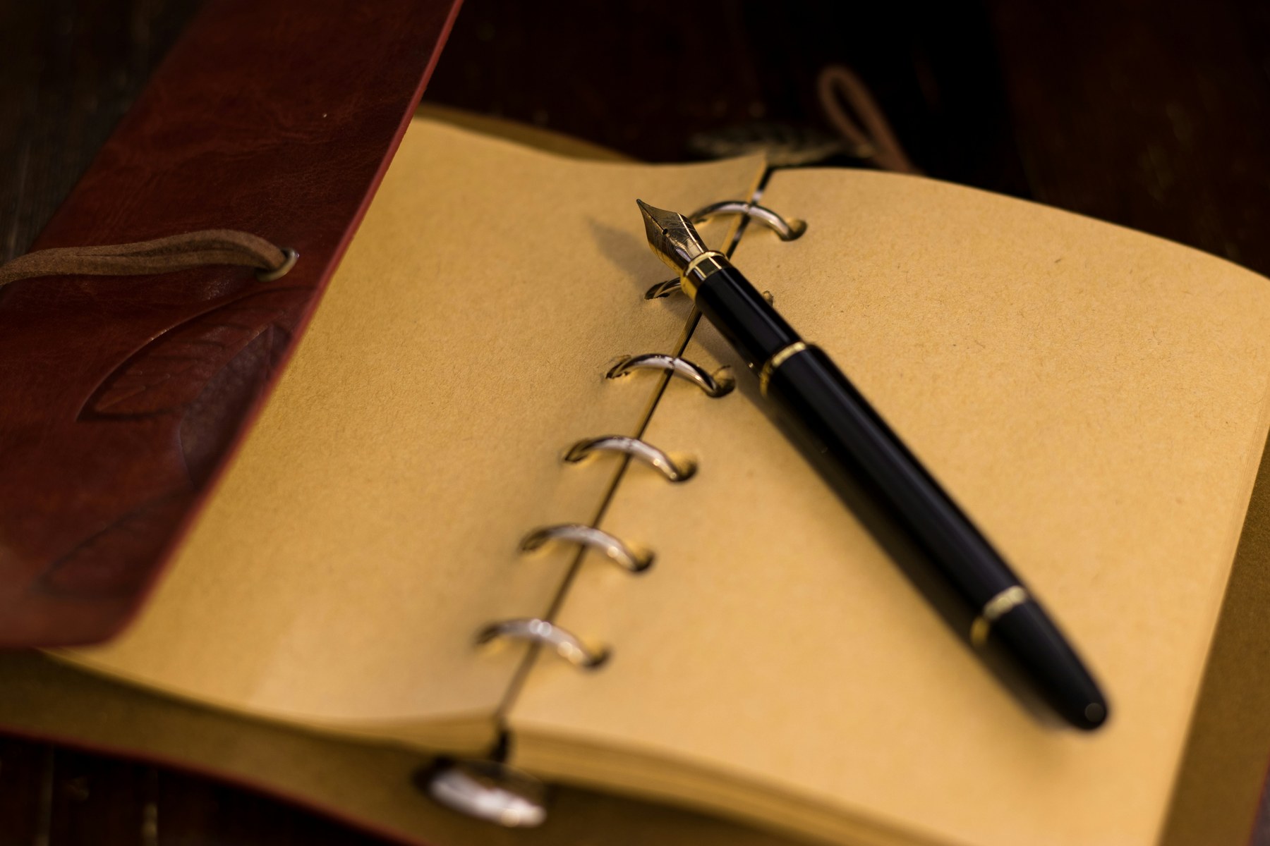 Carnet relié en cuir rouge ouvert sur des pages kraft, stylo-plume noir et plume dorée posé en diagonale — registre de maison.